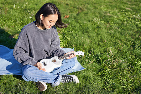 微笑的亚洲女孩 音乐家在公园里演奏四弦琴 在阳光明媚的日子里 小心翼翼地看着她的乐器 坐在毯子上图片