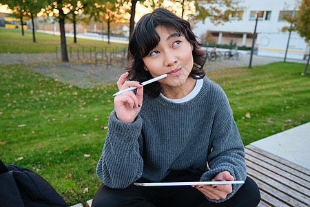 亚洲女性的画像 公园里的学生 坐在长椅上 拿着数字平板电脑和一支笔 思考 一边思考 一边做笔记 在户外做作业女孩快乐自由职业者设图片