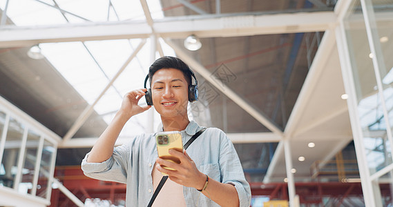 电话 音乐耳机和走在商场流媒体播客或广播中的亚洲人 技术 旅行和快乐的男性在城市建筑的移动智能手机上收听歌曲 音频或播放列表图片