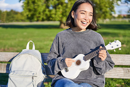 快乐的可爱女孩独自坐在公园的长椅上 弹四轮吉他 在户外享受阳光明媚的一天爱好音乐家成人黑发音乐女性喜悦乐器唱歌乐趣图片