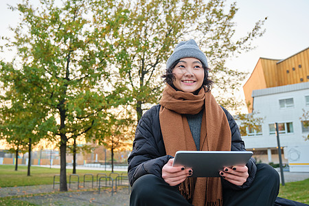 穿着暖和衣服的亚洲女孩的画像 坐在长椅上 拿着数字平板电脑和图形笔 开心地笑着 在寒冷的天气里在户外画画药片街道学生女性笔记用户图片