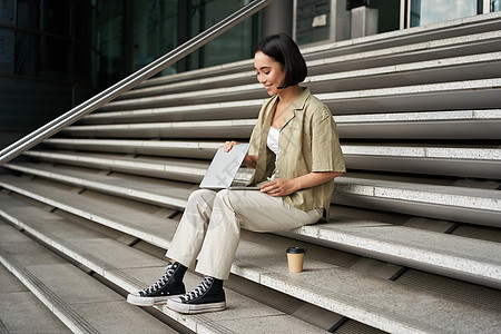 年轻的微笑亚洲女孩带着笔记本电脑 坐在城市的街道上喝咖啡 年轻女性坐在街边的楼梯上用电脑做作业 数字游牧者远程工作快乐电话互联网图片
