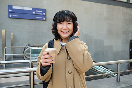 美丽的微笑韩国女孩 喝外卖咖啡 戴着耳机听音乐 穿着风衣和背包 站在火车站 旅行游客旅游音乐车站成人歌曲快乐互联网女士学生图片