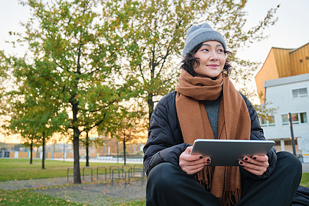 穿着暖和衣服的亚洲女孩的画像 坐在长椅上 拿着数字平板电脑和图形笔 开心地笑着 在寒冷的天气里在户外画画街道自由职业者笔记女士用图片