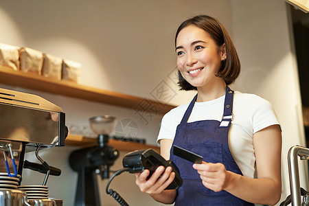 可爱的亚洲女孩 咖啡馆里的咖啡师处理非接触式支付 将信用卡插入 POS 终端 在咖啡店点菜职员酒吧围裙商业机器食物工作卡片咖啡服图片