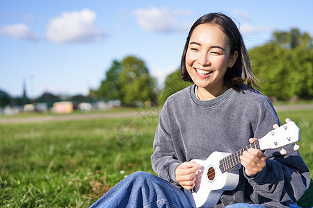 无忧无虑的亚洲女孩在公园里唱歌和弹尤克里里 坐在草地上 音乐家在户外闲暇时放松歌曲弦琴吉他乐趣花园爱好快乐幸福黑发乐器图片