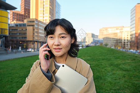 时尚的微笑女孩 穿着风衣的大学生 拿着平板电脑 用手机说话 通过电话交谈 看起来很放松 站在街上药片街道互联网界面设计师快乐成人图片