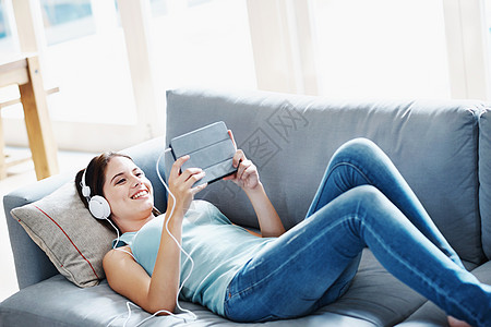 在她的平板电脑上玩耍 一个迷人的年轻女人躺在家里的沙发上 在她的平板电脑上听音乐图片