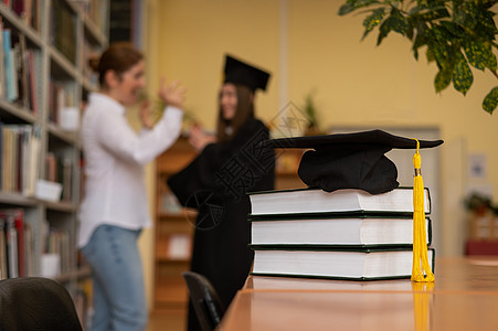 在图书馆的一堆书上加盖了毕业帽 教师和背景中的女青年毕业生都完成了毕业封套学校考试架子班级大学桌子书架学习老师流苏图片