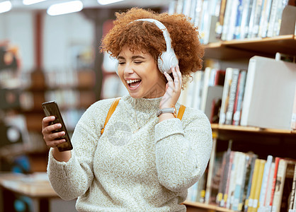 黑人女性 电话和图书馆里的音乐 戴着耳机 在网络应用程序上流媒体和大笑 大学生 有趣的博客或社交媒体 带微笑 智能手机和学习 以图片