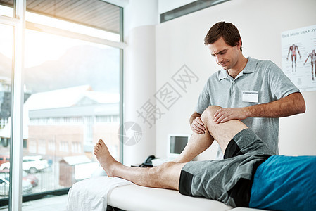 当我们完成后 您的伤害将成为过去 一位年轻的男性物理治疗师帮助躺在床上的客户进行腿部锻炼图片