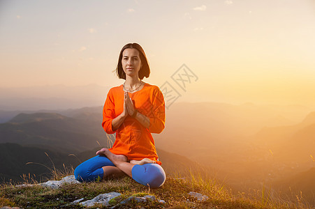 尤吉女人坐在莲花的姿势下 双腿交叉 在山上日落的背景下将手握在胸前 旁边有一个广告场所 (Yeogi女士)图片