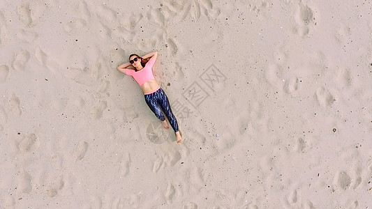 夏天 戴着太阳镜的时尚年轻运动美女躺在沙滩上 在荒凉的海滩上 从上面看 航空视频肌肉微笑自由练习日落身体女孩专注天线训练图片