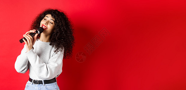 娱乐和爱好概念 时尚的年轻女子唱卡拉OK 仰视并拿着麦克风 表演歌曲 站在红色背景上 笑声购物女孩歌手广告情感促销工作室化妆品嘴图片
