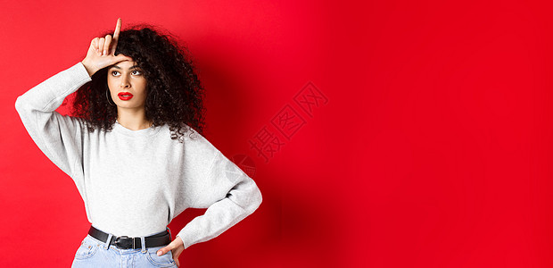 有卷发的自大女人 额头显示失败者标志 向前看一旁 嘲笑某人 站在红背景上女士购物黑发魅力情感工作室生活促销广告卷曲图片