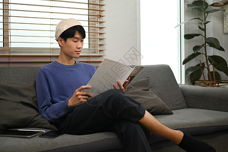 和平的年青亚洲男子在沙发和阅读书上放松 在家闲暇时间图片