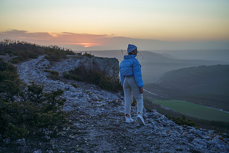女游客在日出山顶上 女孩向太阳致敬 穿着蓝色夹克 白帽子和白色牛仔裤 概念设计石头阳光天空旅游女士自由冒险顶峰悬崖旅行图片