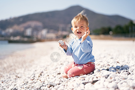 笑着的孩子一只手拿着一块鹅卵石 另一只手举在鹅卵石滩上 背景是群山图片