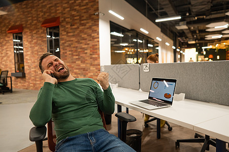 在现代联合办公空间的办公桌前 留着胡子的白种人用智能手机聊天笔记本电脑男人办公室教育影响者网站企业家商务雇主图片