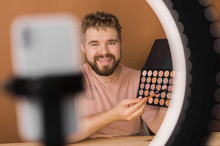 男人在视频博客上谈论化妆和美容产品 拍摄化妆教程的男性 Guy 教授使用她的手机制作教程  多样性和博主概念环形女性刷子营销播送图片