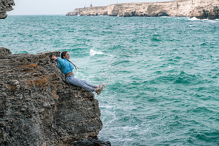 一位身穿蓝色夹克的女子坐在悬崖峭壁上方的岩石上 望着波涛汹涌的大海 女旅行者休息 思考 梦想 享受大自然 和平与平静的景观 多风图片