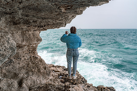 一位身穿蓝色夹克的女子站在悬崖峭壁之上的岩石上 眺望着汹涌的大海 女旅行者休息 思考 梦想 享受大自然 和平与平静的景观 多风的图片