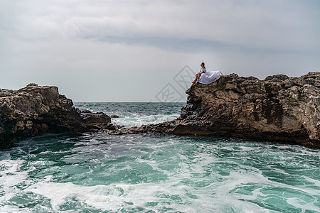 暴风雨中的一个女人坐在海面的一块石头上 穿着白色长裙 波浪冲向岩石 白喷雾上升海浪海洋头发蓝色海岸冲浪碰撞旅行日落悬崖图片