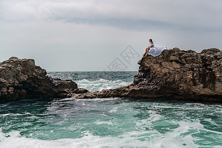 暴风雨中的一个女人坐在海面的一块石头上 穿着白色长裙 波浪冲向岩石 白喷雾上升支撑海景女孩女性悬崖太阳风暴碰撞头发海滩图片