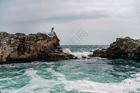 暴风雨中的一个女人坐在海面的一块石头上 穿着白色长裙 波浪冲向岩石 白喷雾上升太阳碰撞支撑海洋女性海景天空头发旅行风暴图片