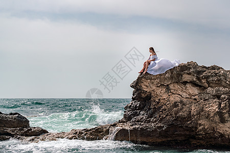暴风雨中的一个女人坐在海面的一块石头上 穿着白色长裙 波浪冲向岩石 白喷雾上升女性天空太阳成人悬崖海洋支撑蓝色海景海浪图片