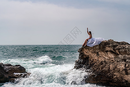 暴风雨中的一个女人坐在海面的一块石头上 穿着白色长裙 波浪冲向岩石 白喷雾上升风暴碰撞支撑女性天空日落太阳成人旅行海浪图片