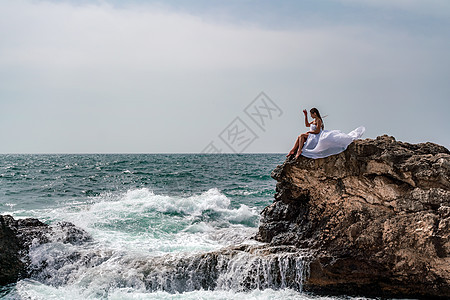 暴风雨中的一个女人坐在海面的一块石头上 穿着白色长裙 波浪冲向岩石 白喷雾上升女士女性碰撞成人海景海浪风暴旅行海岸蓝色图片