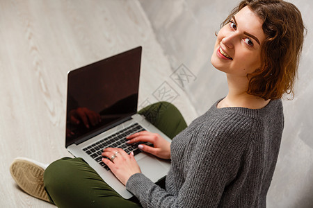 满意的女性画像 带着美丽的微笑 享受着用银色电脑看电影 坐在灰色墙壁上的地板上的莲花姿势幸福工作技术学习女孩学生地面黑发冲浪女士图片