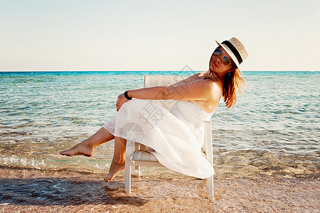 身着白裙子和帽子的年轻女子坐在海中的椅子上 海岸 坐着图片