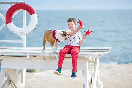 英俊的男孩少年快乐地与他的朋友斗牛犬在海边共度时光 小狗抱着玩两颗海星靠近救生圈漂浮穿着红色裤子拖鞋和 T 恤 运动 宠物图片