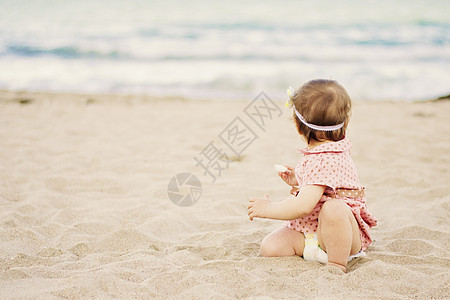 可爱的女婴穿着波尔卡圆点上的衣服 坐在海边沙滩上的沙子上 望着大海图片