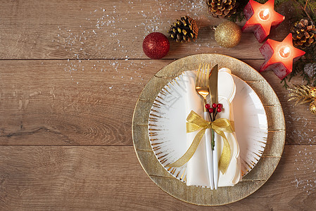 圣诞假期晚餐餐位布置-盘子 餐巾 餐具 橡木桌背景上的金色摆设装饰 金盘子上的叉子和勺子 在红色蜡烛 锥体和球附近 平躺图片