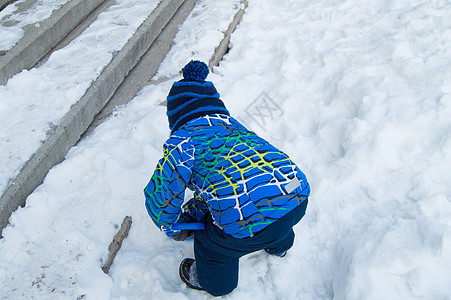 冬天 一个戴着蓝色针织帽和暖和夹克的小男孩在积雪覆盖的楼梯附近玩耍 从后面看 美丽的 天气图片