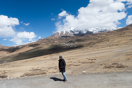 身穿冬衣的印度旅行者独自站在遥远的山路上 背景中 雪峰在山顶上飘着浮云蓝天图片