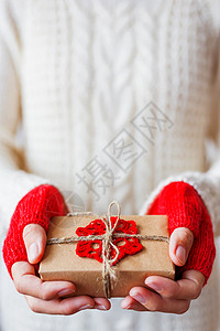 穿着白色针织毛衣和手套的女人拿着一份礼物 礼品采用带有钩编红色雪花的牛皮纸包装 包装礼物的 DIY 方式示例 白色的 粗糙的图片