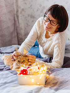 可爱的姜黄色猫 有着由装饰性羽毛制成的亮金色耳朵 女人在玩她毛茸茸的宠物和装有圣诞装饰品的盒子 新年前舒适的家 十二月 毛皮图片