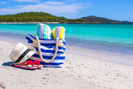 白沙滩上的蓝包 草帽 翻滚式拖鞋和毛巾 太阳镜图片