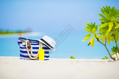 白沙滩上的蓝袋 草帽 墨镜和防晒霜瓶 蓝色的背景图片