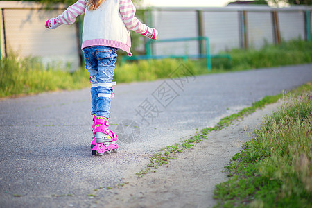 拎着轮滑鞋夏天 一个小女孩在沥青上骑着粉红色溜冰鞋 这孩子学会了骑滑雪鞋 户外活动 童年背景