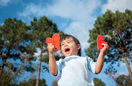 可爱的小女孩拿着两个红色心形回收纸板在蓝天和绿树背景上 情人节的爱情象征 爱情和浪漫的概念 户外 假期图片