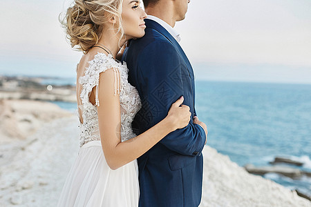 在亚得里亚海沿岸的白色岩石上 身穿白色蕾丝连衣裙的年轻金发美女倚在身穿时尚蓝色西装的英俊男子身上 穿着婚纱的模特女孩和时髦的新郎图片
