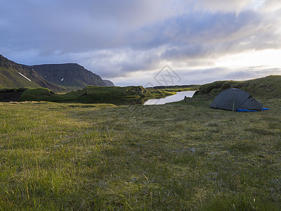 小蓝色帐篷独自站在冰岛长满苔藓的溪岸上的绿草上 积雪覆盖的山丘和悬崖 多云的天空背景 黄金时段的灯光 复制空间 河 远足图片