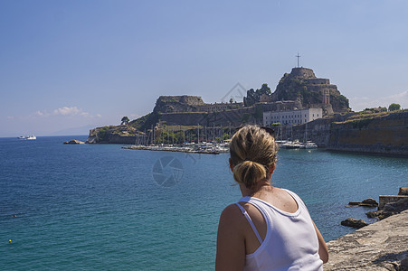 年轻金发金发美女在著名的旅游标志式的老威尼斯堡垒上观视 时钟塔和夜总会港 克尔基拉市 希腊科孚 夏日 海面和蓝天空背景图片