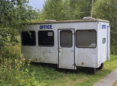 小型白色拖车小屋 配有蓝色标志办公室 在人行道附近农村地貌的废弃大篷车 绿草和树木 金属 沙漠图片