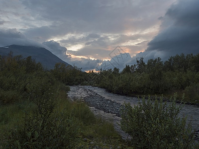 与白桦树森林和山脉在夕阳光下的野生河流 夏季的拉普兰自然景观 喜怒无常的天空 粉红色的云彩图片